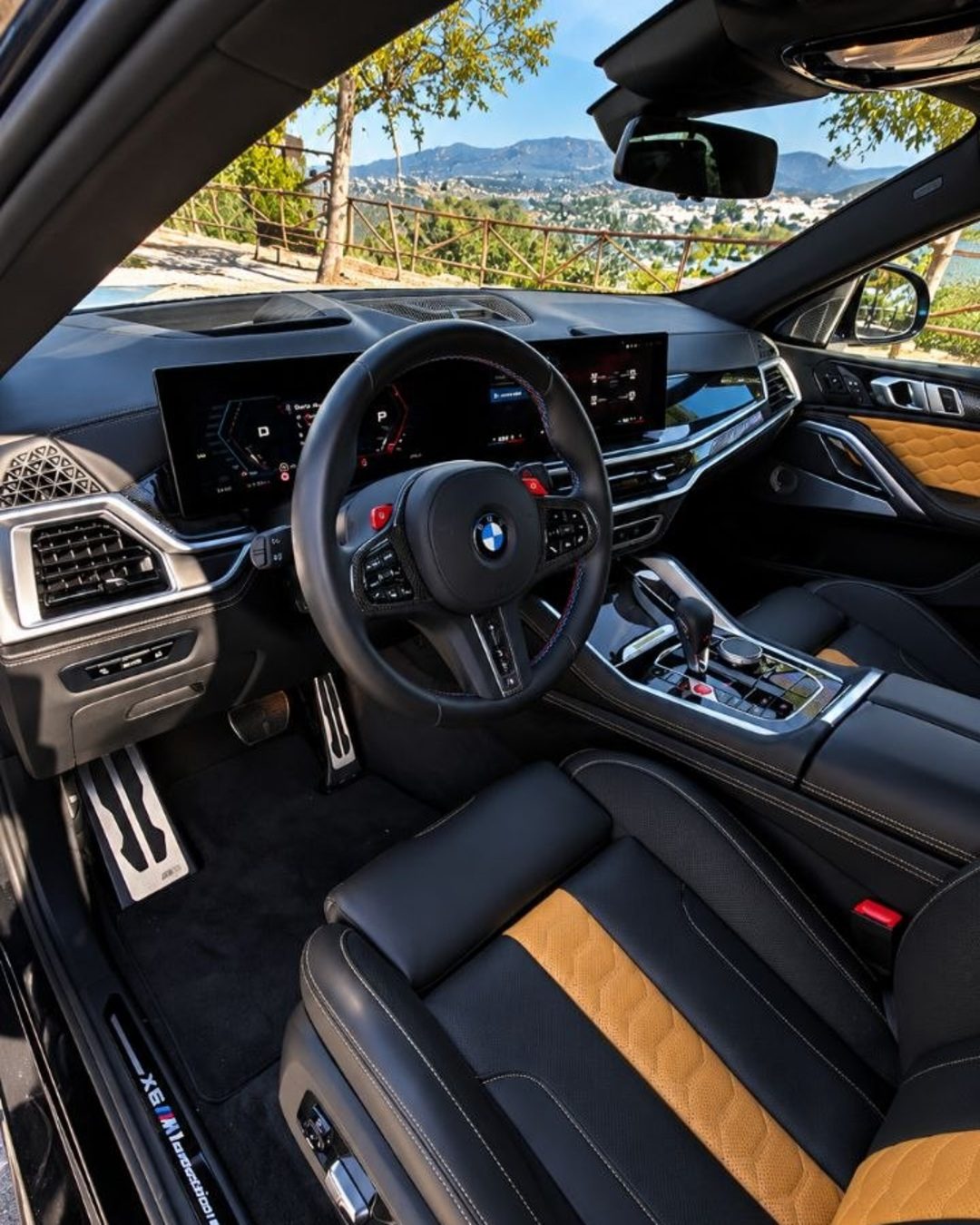 Водительское место BMW X6 M с рулём M, цифровыми дисплеями, центральной консолью и чёрным кожаным салоном.