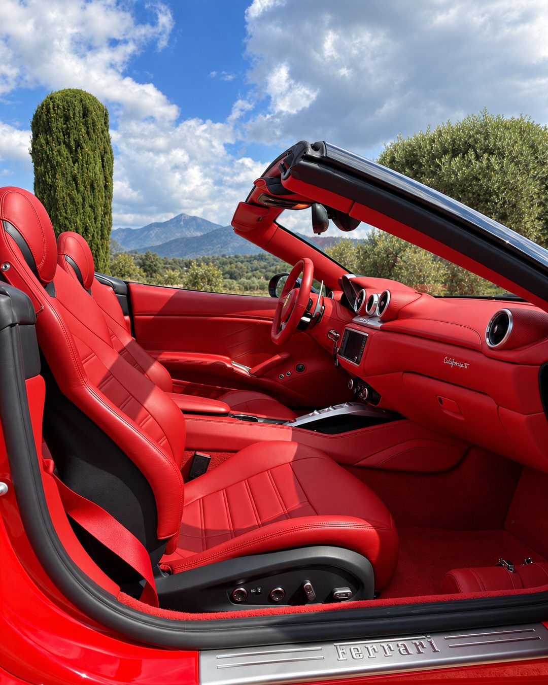Ferrari California T - Red leather seats and dashboard of the Ferrari California T seen from the passenger side with the roof open, mountains and blue sky behind.